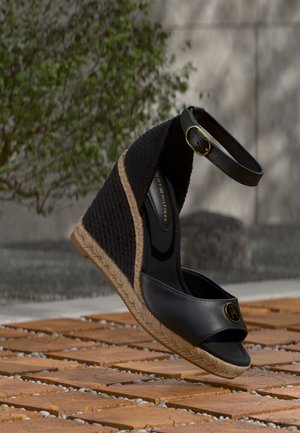 Sandalo zeppa nero con cinturino alla caviglia, dettaglio in corda sulla suola e logo Tommy Hilfiger sul davanti, posizionato su un pavimento piastrellato all'aperto.