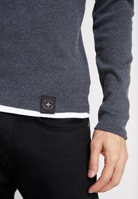 Gray long-sleeve sweater with ribbed texture, featuring a black fabric tag at the hem. Worn over a white shirt and paired with black jeans.