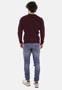 Uomo con capelli ricci che indossa un maglione a maglia bordeaux, jeans blu e sneakers bianche, in piedi di spalle su uno sfondo neutro.