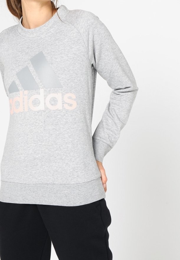 Femme portant un sweat-shirt Adidas gris clair avec logo et un pantalon noir, debout avec une main derrière le dos.