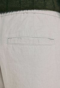 Gros plan sur la poche arrière d'un pantalon en lin beige avec taille élastique et haut vert foncé rentré à l'intérieur.