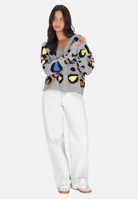 Cardigan grigio con macchie leopardate multicolori in giallo, rosa, blu e nero, con scollo a V e chiusura con bottoni, indossato su jeans bianchi.