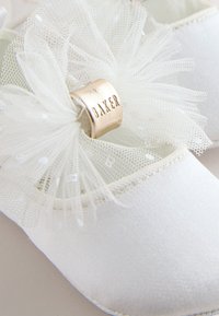 Scarpe in tessuto bianco con un dettaglio floreale in tulle e un'etichetta metallica dorata con la scritta "DAKER". Texture liscia con decorazioni delicate.