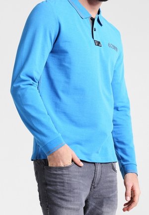 Homme portant un polo à manches longues bleu vif avec col boutonné et jeans gris foncé, main dans la poche, devant un fond uni.