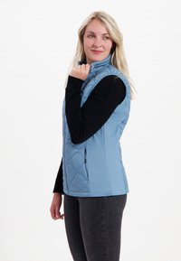 Ljust blå isolerad väst med hög krage, kviltat mönster och dragkedjefickor, kombinerad med en svart långärmad topp och mörka jeans.