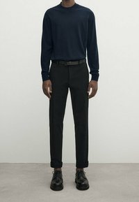 Marineblauer Langarmpullover, schwarze Hose und schwarze Lederschuhe. Der Pullover hat einen Rundhalsausschnitt; die Hose hat einen geraden Schnitt und hochgekrempelte Säume.