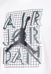 T-shirt en coton blanc avec un graphique "AIR JORDAN" en noir et gris, avec la silhouette d'une figure en train de sauter et un motif en grille en arrière-plan.