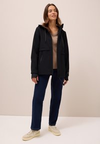 Zwarte softshell jack met capuchon, twee voorzakken en trekkoordverstelling. Gepaard met een beige top en donkerblauwe rechte broek.