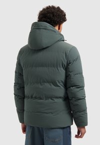 Man in een groene gewatteerde jas met capuchon, gezien vanaf de achterkant, gecombineerd met blauwe denim jeans tegen een effen achtergrond.