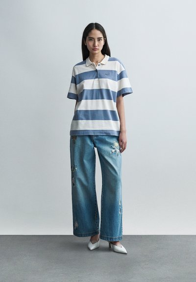 Ριγέ πόλο πουκάμισο σε ανοιχτό γαλάζιο και λευκό, συνδυασμένο με φαρδιά τζιν παντελόνια με κεντήματα. Το μοντέλο φοράει άσπρες γόβες, παρουσιάζοντας ένα casual σύνολο.