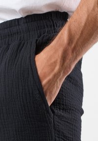 Sorte teksturerede bukser med elastisk talje og sidelomme, der fremviser en krøllet stoffinish. Mærket er synligt nær lommen.