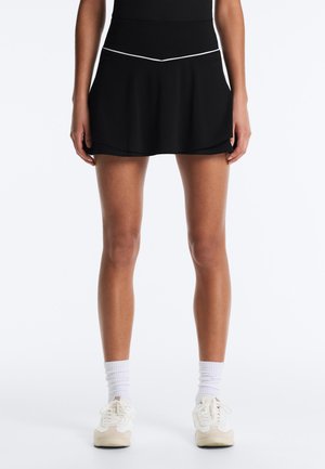 Fekete sport szoknyát fehér díszítéssel viselő személy alsó teste, fehér boka zoknival és fehér tornacipőkkel egyszerű háttér előtt.