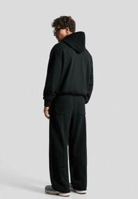 Felpa nera con cappuccio abbinata a pantaloni larghi coordinati. Tessuto morbido, tasche laterali e cuciture minime. Modello che indossa sneaker grigie.