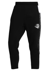 Černé tapered tepláky s elastickým pasem, skvrnitými bočními panely, bílými pruhy a emblémem lebky s křídly na horní části stehna.