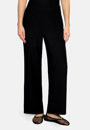 Vrouw draagt zwarte broek met wijde pijpen en zwarte mesh ballerina's, staand voor een effen lichte achtergrond.