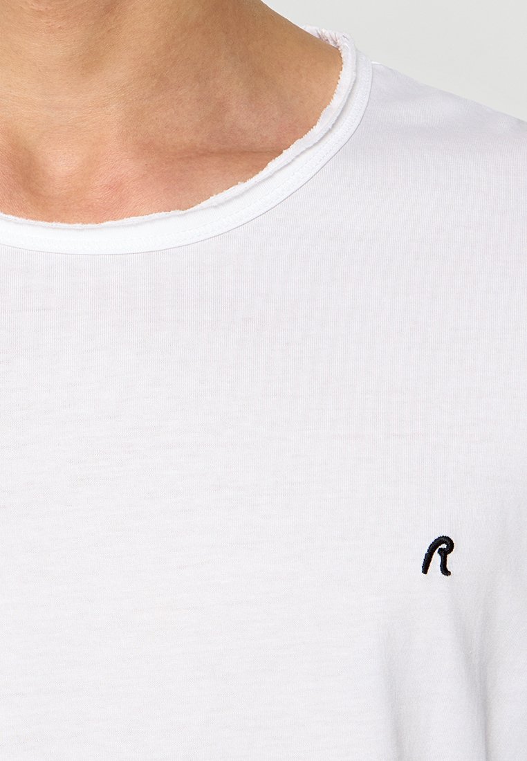 Primer plano del cuello y la parte superior del pecho que lleva una camiseta blanca de cuello redondo con un pequeño logo bordado en negro con una "R" en el lado derecho.