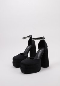 Zapatos de plataforma negros con un alto tacón cuadrado, hechos de un material similar a ante, que cuentan con una correa en el tobillo adornada con cristales y un diseño de punta redonda.