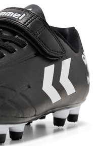 Crampon de football noir avec une tige en synthétique texturé, comportant des accents en chevron blancs et une sangle Velcro. Équipé de crampons noirs et blancs.