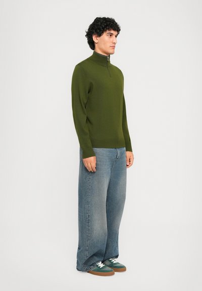 Mann med krøllete mørkt hår iført en olivengrønn glidelåsgenser, vide falmede blå jeans og grønne og hvite joggesko som står sidelengs.