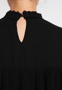 Černé šaty s texturovaným krajkovým límcem a detailem ve tvaru klíčové dírky na zádech, s skládáním látky sahajícím od pasu.
