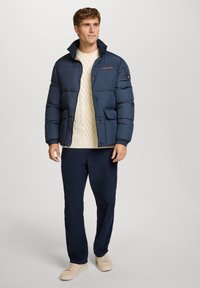 Giacca puffer blu marina con colletto alto, chiusura a bottoni e due tasche frontali, abbinata a un maglione crema a trecce e pantaloni scuri.