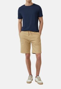 Marineblaues Kurzarm-Chemisier aus Baumwolle kombiniert mit hellbeigen Shorts mit Kordelzug. Das Modell trägt weiße Sneaker mit beigen Akzenten. Lässiges Outfit.