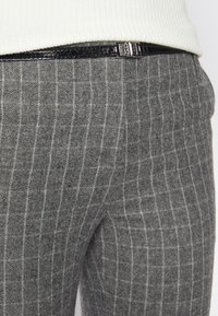 Gros plan d'un pantalon gris à fines rayures avec une fine ceinture noire brillante et une partie d'un haut blanc côtelé visible à la taille.