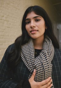 Black checkered coat with a blue and beige houndstooth scarf. Long dark hair, holding the scarf with a hand resting on it.