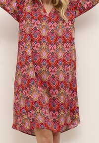 Kvinna klär i en knähög klänning med långa ärmar som har ett livligt mönster i rött, rosa, guld och blått med geometriska och blommönster.