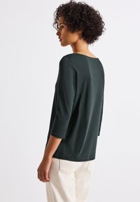 Maglietta verde scuro a tre quarti, con un taglio rilassato e scollatura ampia. Tessuto liscio senza motivi visibili o dettagli metallici.