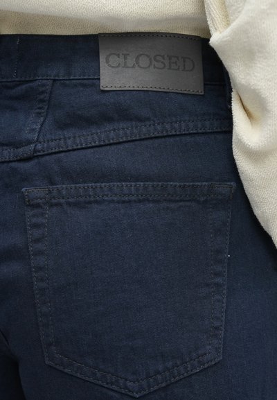 Tumši zili džinsa džinsi ar taisnstūrveida kabatu un izšūtu apmali. Jostasvietā ir melna ādas plāksnīte ar uzrakstu "CLOSED".