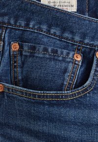 Blå denimjeansficka med kopparnitar och synlig orange sömnad, med en liten inre myntficka och en delvis synlig varumärkesetikett.