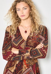 Langärmliges, gemustertes Kleid mit tiefem V-Ausschnitt, in satten Rot-, Schwarz- und Goldtönen, akzentuiert durch Knopfdetails und florale Muster.