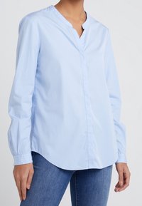 Jasnoniebieska koszula z długim rękawem, uszyta z gładkiego materiału, z dekoltem w kształcie litery V, bez guzików z przodu oraz zaokrąglonym dołem. Luźny krój.