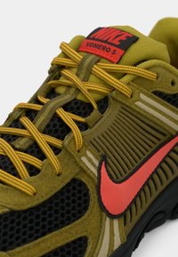 Olivovo zelené a čierne tenisky Nike Vomero 5 s oranžovými šnúrkami a červeným logom Nike na boku a jazyku.