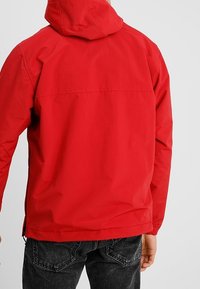 Red hooded jacket with a smooth, water-resistant fabric. Features a rear pocket seam and adjustable hem for a snug fit.