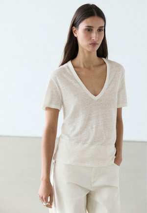 Femme aux longs cheveux bruns portant un haut à manches courtes beige clair avec un col en V et un pantalon assorti de couleur claire, debout devant un fond uni.