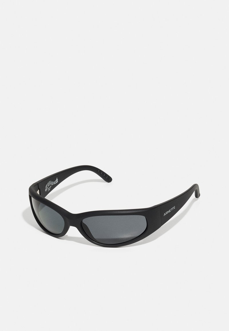 Zwarte zonnebril met een gebogen montuur en donkere lenzen. Beschikt over een matte afwerking en de merknaam "ARNETTE" aan de zijkant.