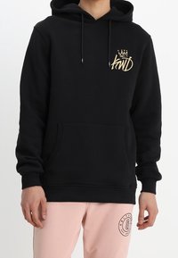 Person som bär en svart hoodie med guldkronslogotyp på bröstet och ljusrosa byxor med cirkulärt emblem på vänstra låret.
