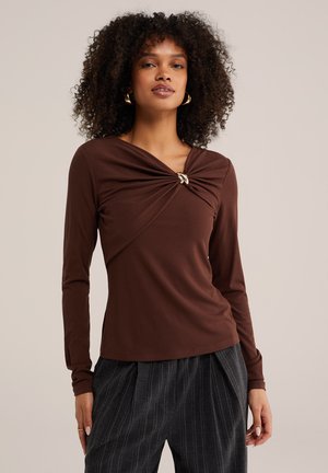 Femme aux cheveux bouclés portant des boucles d'oreilles anneaux dorés, un haut marron à manches longues avec un détail torsadé sur le devant, et un pantalon sombre à fines rayures.