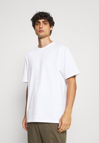 Camiseta de algodón blanca con mangas cortas, cuello redondo y corte relajado, combinada con pantalones verde oliva. Textura suave, sin patrones visibles.