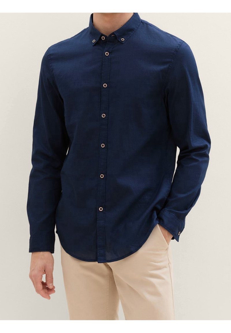Homme portant une chemise ajustée bleu marine à boutons avec un pantalon beige, une main dans la poche, debout devant un fond clair uni.