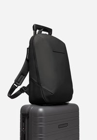 Sac à dos noir avec une texture lisse et des bretelles rembourrées, reposant sur une valise rigide grise. Comprend une poche avant et un design épuré.