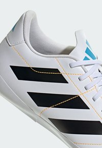 Hvide sportsko med sorte striber, der har teksturerede materialer, orange syninger og lyseblå detaljer på hælen og logoet.