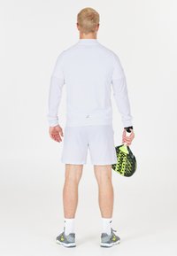 Hvid langærmet bluse og shorts, lavet af let stof. Holder en sort og gul paddel. Grå sportssko med hvide sokker.