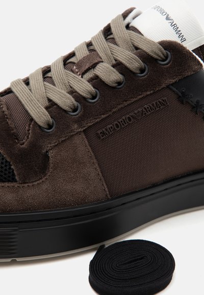 Κοντινό πλάνο καφέ αθλητικού παπουτσιού Emporio Armani με υφή υφάσματος, λεπτομέρειες από σουέτ, γκρι κορδόνια, μαύρη σόλα και ένα επιπλέον ζευγάρι μαύρα κορδόνια δίπλα.