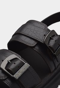 Κοντινό πλάνο σκούρου καφέ δερμάτινου σανδαλιού με υφή, με δύο ρυθμιζόμενες μεταλλικές αγκράφες και παχύ πέλμα.