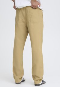 Pantaloni in cotone kaki a vestibilità larga con due tasche posteriori, gambe dritte e texture minima. Indossati con calzature a righe bianche e nere.