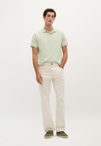 Camisa polo verde claro, pantalones crema, hombre de pie. Material de algodón, mangas cortas, cuello clásico, corte relajado, diseño minimalista, calzado casual.