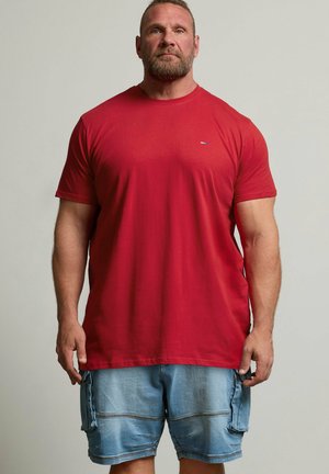 Gespierde man met kort haar en baard, die een effen rood T-shirt en lichtblauwe denim cargoshorts draagt, staand tegen een effen achtergrond.